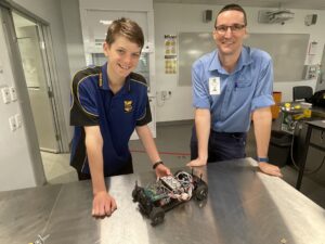 Image of GAWB employee (man) standing to the right of a student from Calliope State School. In front of them on a desk is a model car, fueled by hydrogen. 