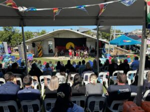 An image of a large crowd of people standing outside in the sunshine watching Darumbal NAIDOC week celebrations.