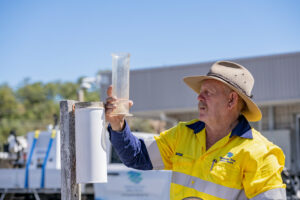 A man in a yellow high visibility shirt and akubra hat is outside, holding up a plastic rain gauge and looking at the amount of water in the gauge.